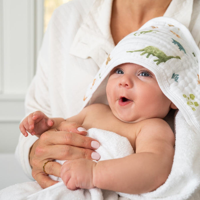 A smiling baby is wrapped in an aden + anais essentials cotton muslin hooded towel featuring dinosaur prints, held by an adult in a white shirt. The softly lit background captures a warm, cozy atmosphere.
