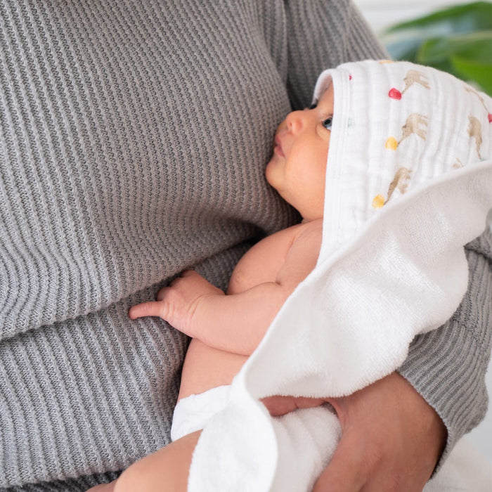 A baby cozily wrapped in an aden + anais essentials cotton muslin hooded towel is held by a person in a gray knit sweater, as the baby gazes upwards.