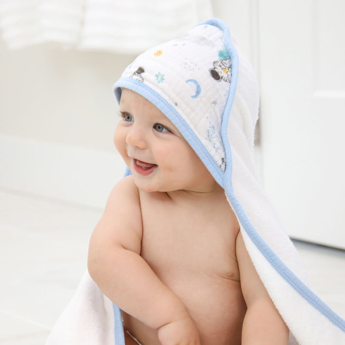 A baby with blue eyes sits on the floor, wrapped in an aden + anais essentials cotton muslin hooded towel. The towel is adorned with playful patterns and has light blue trim, keeping the child cozy and content in a bright, white room.