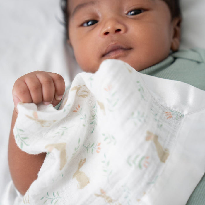 A baby in a green outfit lies calmly on a bed, wrapped in an aden + anais essentials 100% cotton muslin security blanket from a 2-pack, featuring pastel rabbits and leaves. This comforting and stylish setting is ideal for newborns and toddlers.