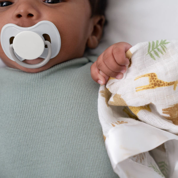 A baby in a mint green outfit, with a gray pacifier, grips the gentle comfort of an aden + anais essentials cotton muslin security blanket, adorned with giraffes and leaves. The focus highlights the babys hand on this soft blanket.