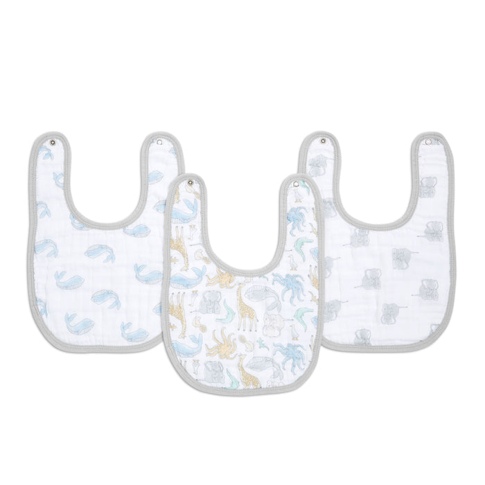 The aden + anais essentials cotton muslin snap bibs 3-pack features absorbent bibs with gray trim: a blue whale pattern on the left, dinosaurs in blue, yellow, and green in the center, and gray mice on the right. These bibs adjust easily as your baby grows.