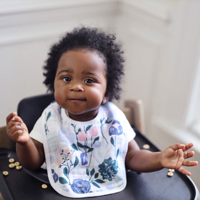 A curly-haired baby with chubby cheeks sits in a high chair, wearing an aden + anais essentials cotton muslin snap bib. They smile slightly in the softly lit, white background, creating a calm ambiance.