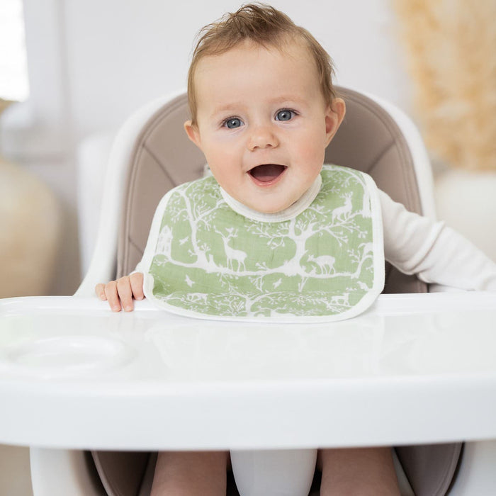 A smiling baby sits in a white highchair, wearing an aden + anais essentials cotton muslin snap bib with white animal and tree patterns. The softly lit room has neutral tones, creating a peaceful setting as the bibs grow with your babe.