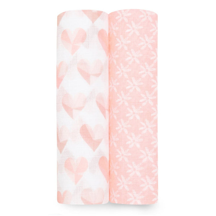 A set of two aden + anais essentials cotton muslin swaddle blankets stands against a white backdrop. One roll features a pink and white heart pattern, while the other showcases a floral design.