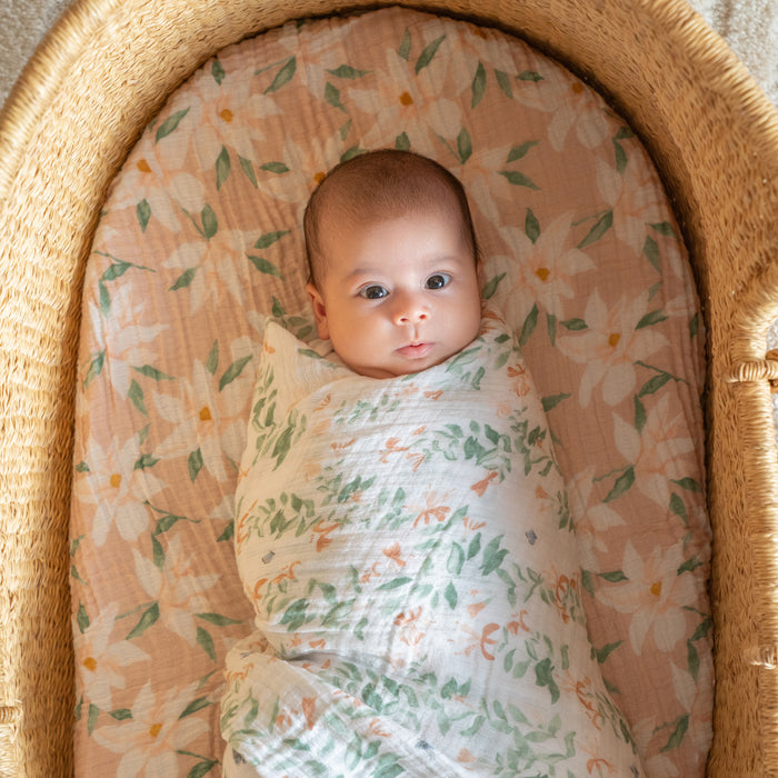 A baby lies in a wicker basket wrapped in an aden + anais essentials cotton muslin swaddle blanket featuring a floral pattern, with a matching background. This versatile product keeps the baby cozy as they gaze wide-eyed at the camera.