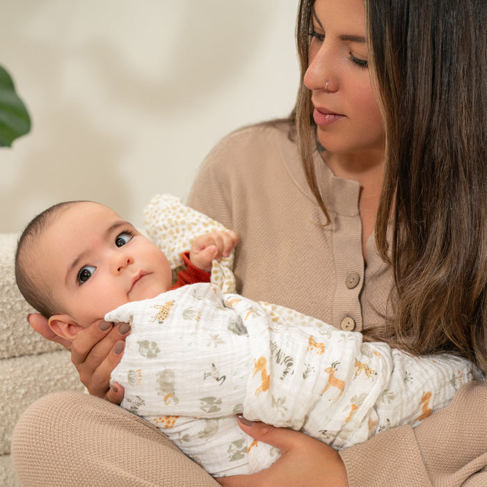 A woman with long hair gazes at a baby wrapped snugly in an aden + anais essentials cotton muslin swaddle blanket, adorned with animal prints, as the baby looks content and towards the camera. The woman is wearing a beige top.
