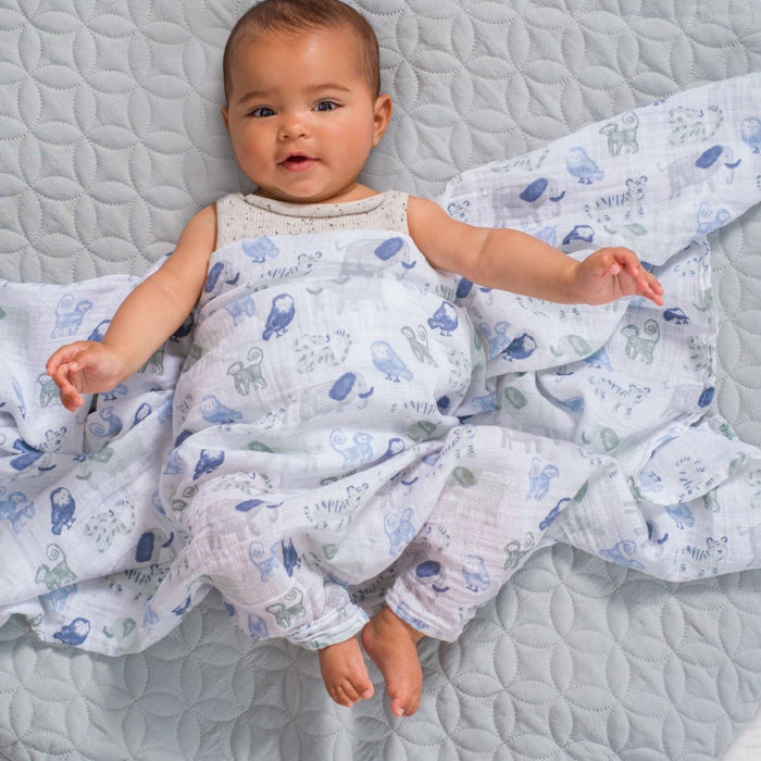 A baby lies on a quilted gray blanket, wrapped in an aden + anais essentials cotton muslin swaddle featuring blue and green animal prints. The cozy fabric keeps the baby smiling as their arms and legs stretch out playfully.