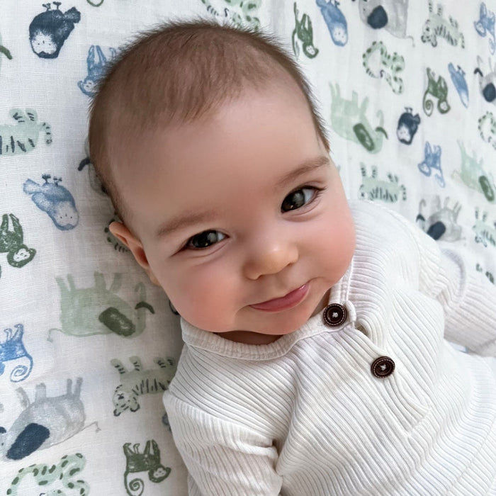 A baby in a white outfit smiles slightly while lying on an Essentials Cotton Muslin Swaddle by aden + anais, featuring cute animal illustrations in green, blue, and gray.