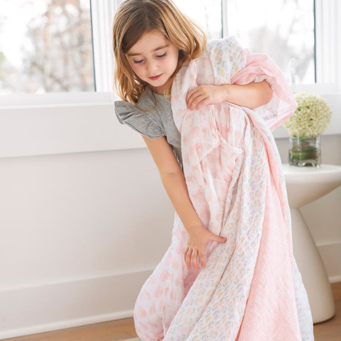 A young girl in a gray top holds an aden + anais essentials cotton muslin swaddle, featuring pink and white patterns. She stands indoors by a window with light curtains, while a small white table with a potted plant sits in the background.