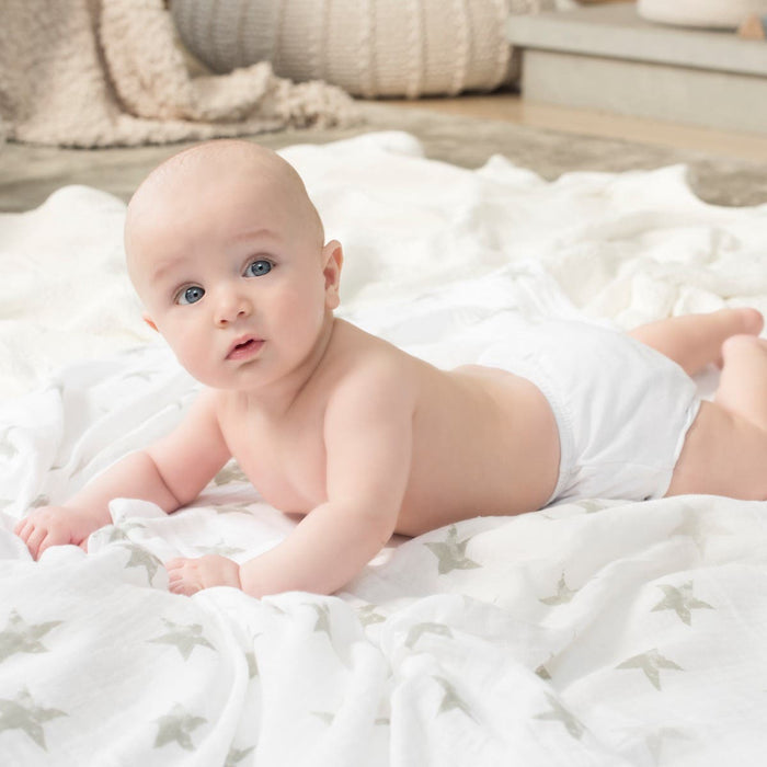 A baby in a white diaper lies on a gray-star-patterned blanket, surrounded by cozy neutral decor, snuggling near aden + anais essentials cotton muslin swaddle blankets.