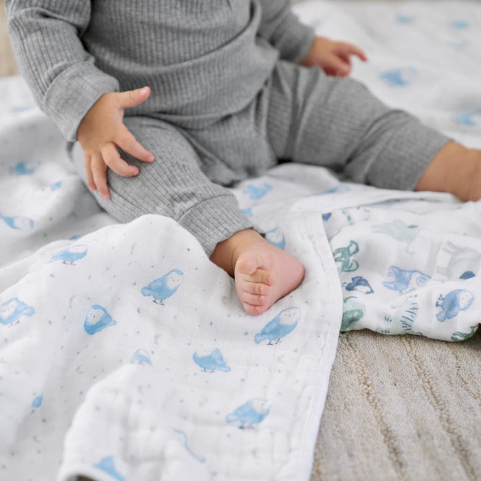 A baby in a gray outfit sits on a white blanket with blue animal prints. The babys legs rest on aden + anais essentials cotton muslin swaddle blanket, while the background shows a textured beige surface.