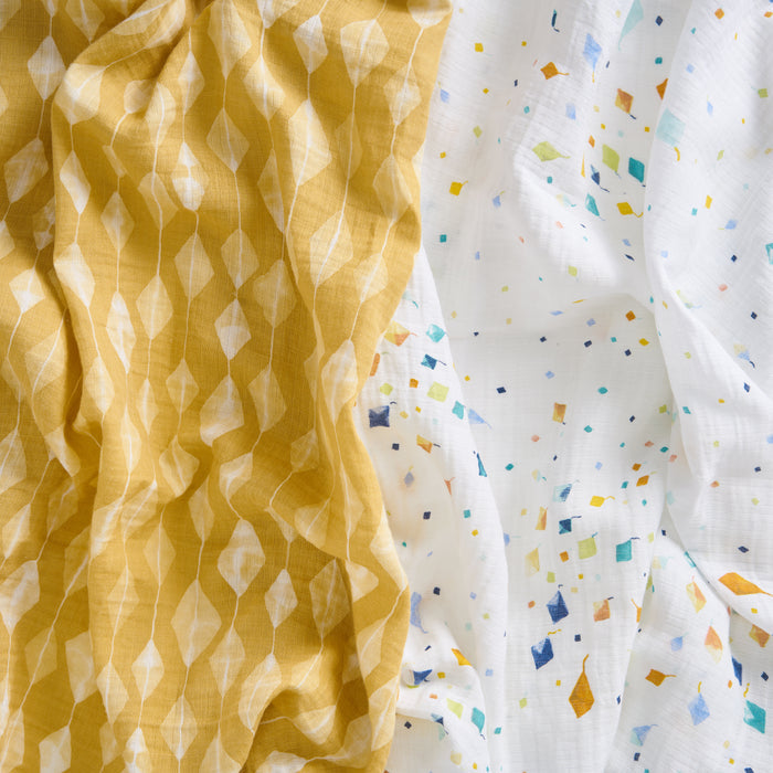 The aden + anais essentials cotton muslin swaddle blanket 2 pack includes a yellow blanket with a white diamond pattern and a white one with a confetti-like square and rectangle design—both light, airy, and perfect for swaddling.