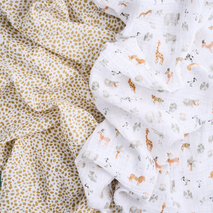 Two pieces of crumpled fabric lie side by side. The left, with a tan speckled pattern, and the right, featuring scattered illustrations of elephants, giraffes, and zebras on white. These breathable swaddles are from the aden + anais essentials cotton muslin swaddle blanket 2 pack line.