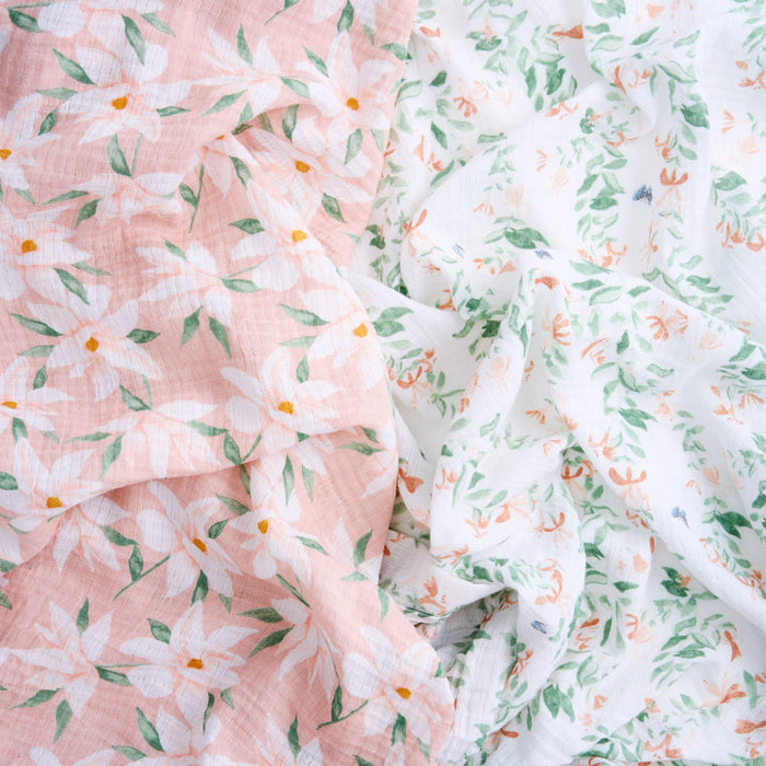 A close-up of aden + anais essentials cotton muslin swaddle blanket shows floral patterns: left side with a pink background, large white flowers, and green leaves; right side with a white background and small blooms in green, orange, and blue—highlighting its versatile use.