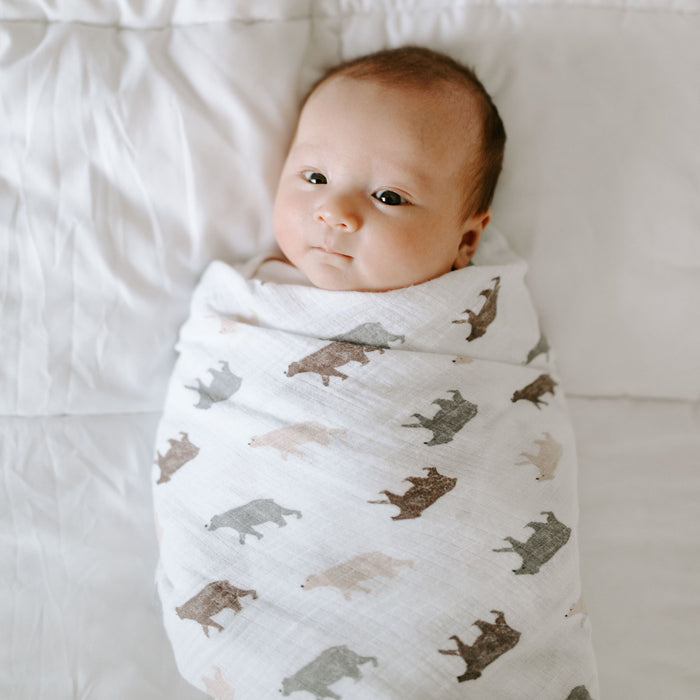 A baby lies calmly on a white bed, wrapped in an aden + anais essentials cotton muslin swaddle blanket featuring animal silhouettes.