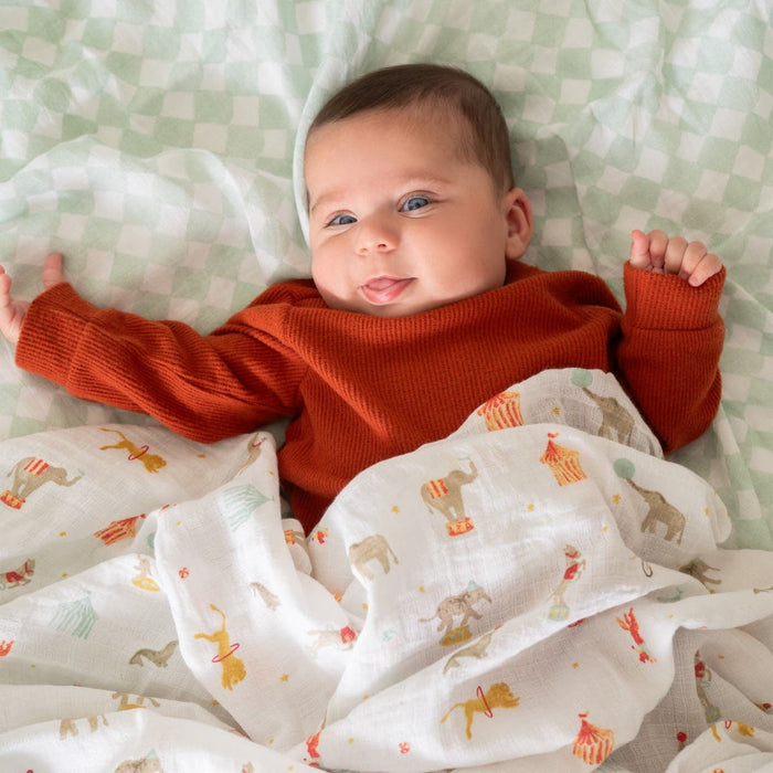 A baby wearing a rust-colored top lies on a soft, checkered blanket, partially wrapped in an aden + anais essentials cotton muslin swaddle from the 4-pack, featuring whimsical animal illustrations like camels and elephants, while smiling contentedly.