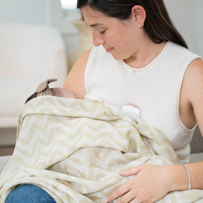 A woman in a sleeveless top cradles a baby wrapped in an aden + anais essentials cotton muslin swaddle blanket. The baby, facing her with a bow on their head, exudes calmness as they sit together on a cozy sofa in softly lit surroundings.