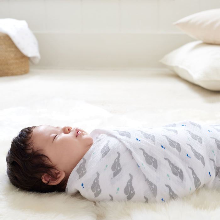 A baby sleeps on a fluffy white rug, wrapped in an aden + anais essentials cotton muslin swaddle blanket featuring gray elephant patterns. The backdrop reveals a light, airy room with minimalist decor, including cushions and a wicker basket.