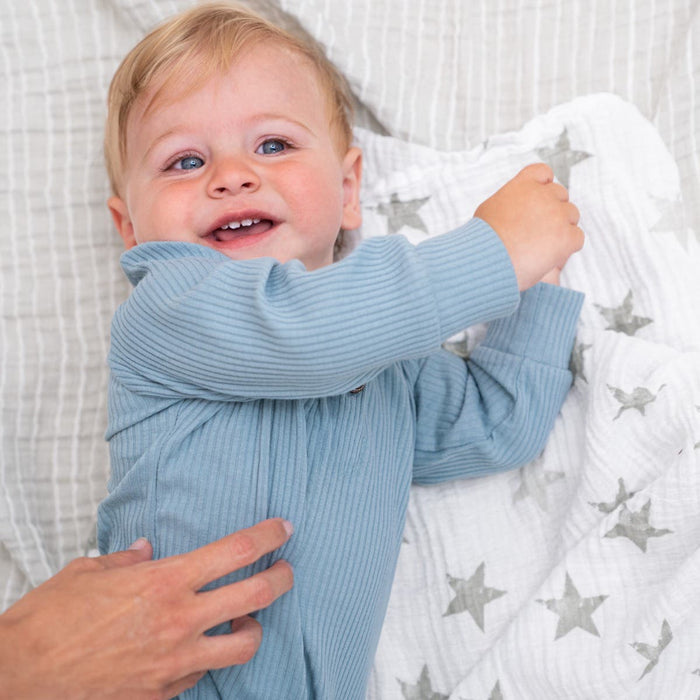 A smiling baby with blond hair lies on an aden + anais essentials cotton muslin swaddle blanket, featuring a star pattern. Wearing a blue ribbed onesie, the baby reaches out as an adults hand gently touches their side.