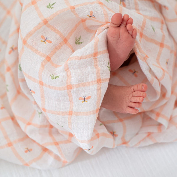 Tiny baby feet peek out from a soft, white aden + anais essentials cotton muslin swaddle blanket featuring orange plaid and butterfly designs, creating a cozy atmosphere of ultimate comfort.