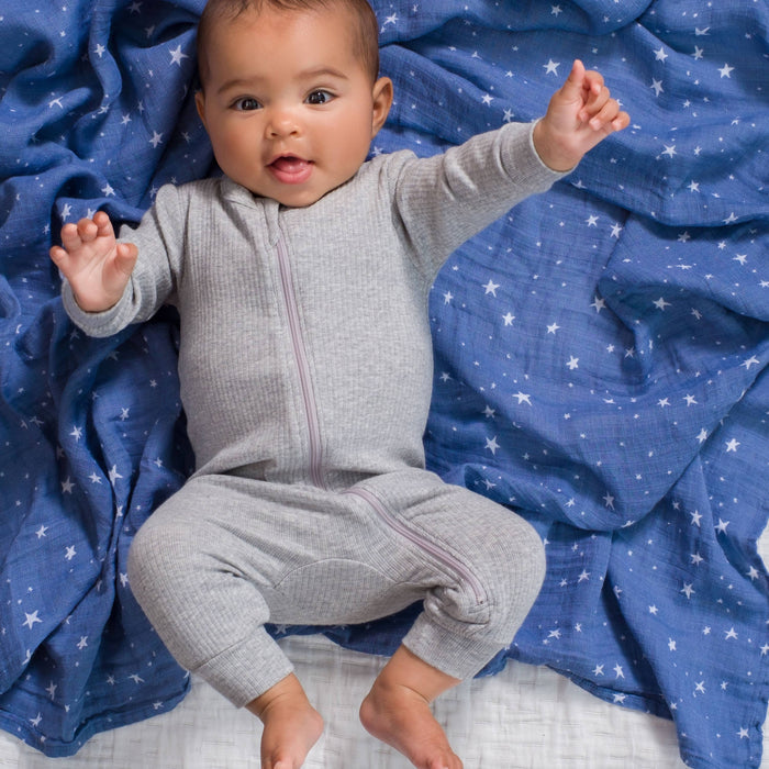 A baby, wearing a gray onesie, is lying on a blue blanket adorned with white stars. The little one smiles and reaches upward, legs bent and arms spread out as if dreaming of the versatile comfort of aden + anais essentials cotton muslin swaddle blankets.
