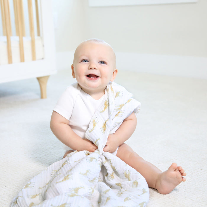 A baby smiles in a white onesie on a light carpet, holding an aden + anais essentials cotton muslin swaddle blanket. In the background, a wooden crib contributes to the rooms bright and airy atmosphere.