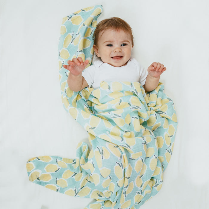 A baby in a white onesie lies on its back, smiling, wrapped in an aden + anais essentials cotton muslin swaddle blanket featuring a yellow and blue leaf pattern. The cheerful scene is set against a bright white background.