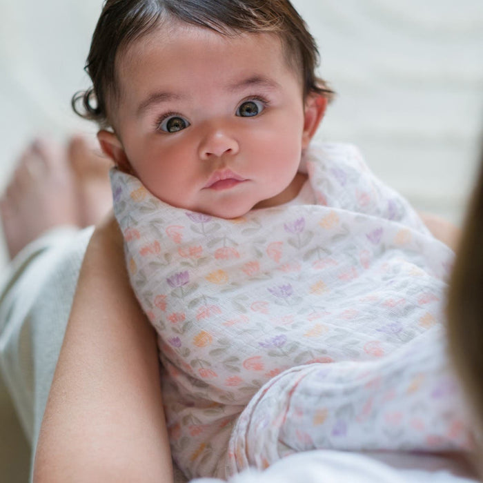 A baby wrapped in an aden + anais essentials cotton muslin swaddle from the 4 pack looks up wide-eyed, with dark hair, cradled by an adult. The breathable fabric ensures comfort as a blurred background highlights the babys expression.