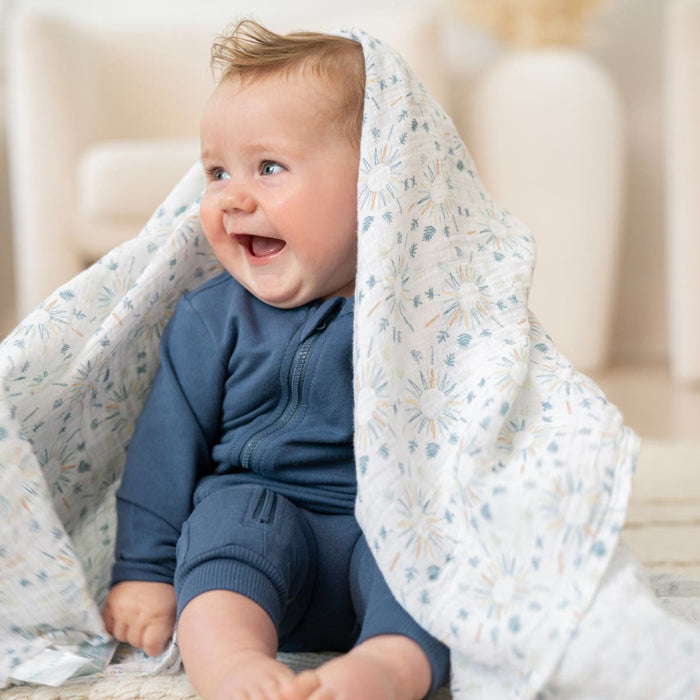 A joyful baby in a blue onesie is on the floor with an aden + anais essentials cotton muslin swaddle draped over their head. The baby smiles broadly, radiating excitement, while the softly lit and blurred background creates a cozy atmosphere.