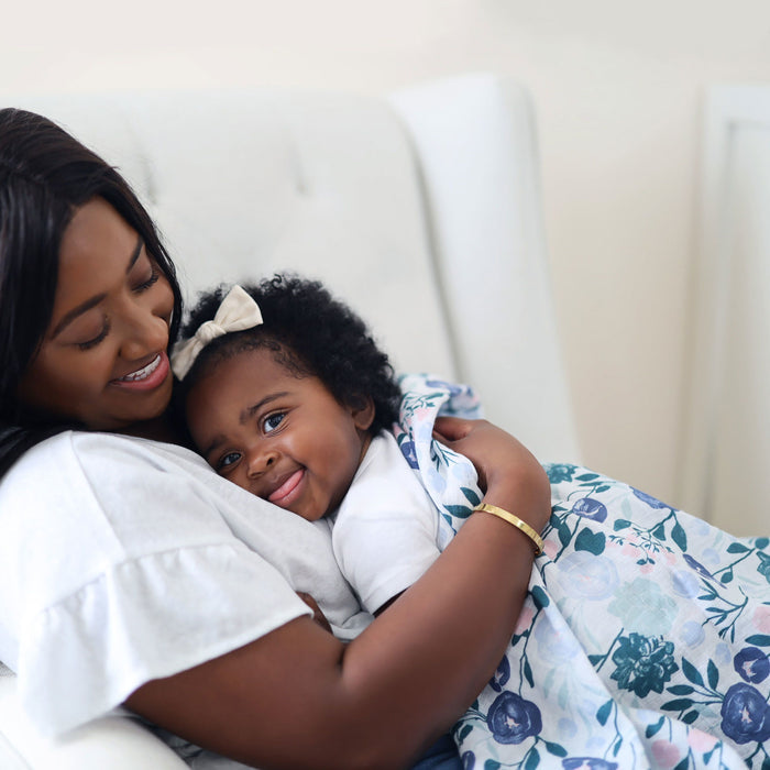 A woman lovingly holds a smiling baby wrapped in an aden + anais essentials cotton muslin swaddle blanket. The baby, adorned with a white bow in her curly hair, gazes up contentedly in the womans embrace. They sit together in a light chair against a soft backdrop.