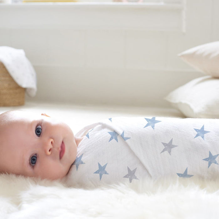 An infant is snugly wrapped in an aden + anais essentials cotton muslin swaddle blanket featuring blue stars, lying on a plush white surface. The softly lit room includes pillows and a woven basket in the background, enhancing the serene ambiance.