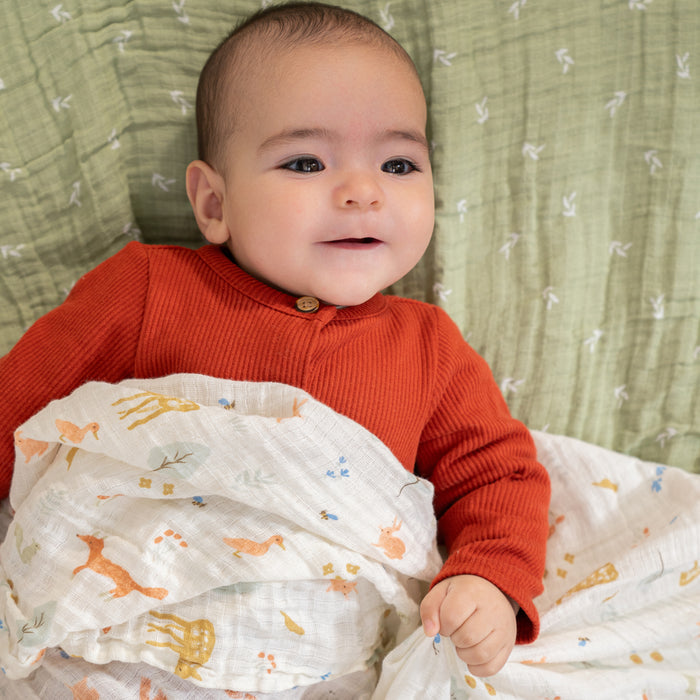 A baby in a red sweater lies on a green blanket, holding an aden + anais essentials cotton muslin swaddle from a 4 pack. The swaddle features colorful animals like giraffes and birds on breathable fabric, gently cradling the little one who gazes up with a soft expression.