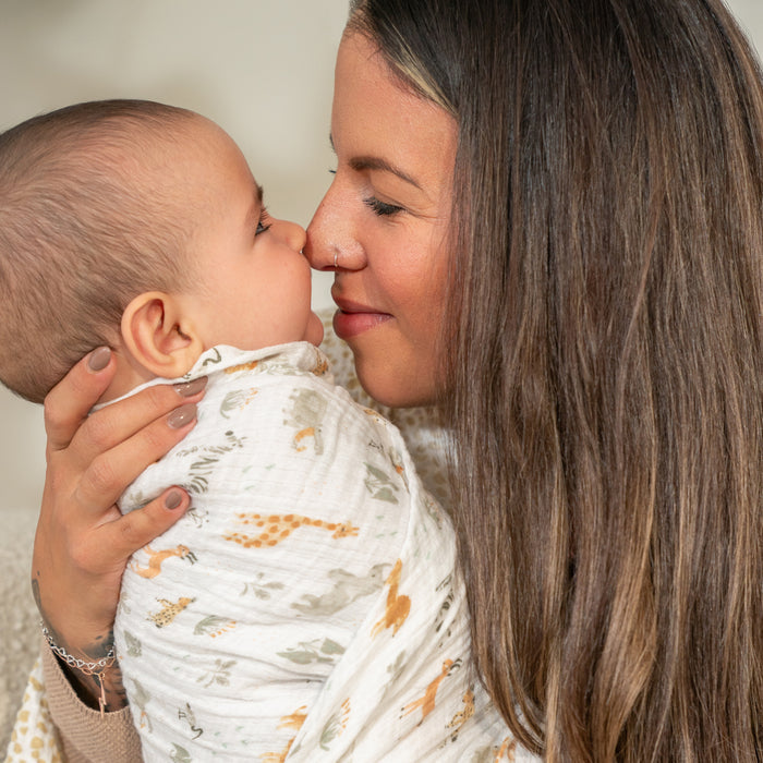 A long-haired woman lovingly touches noses with her baby wrapped in an aden + anais essentials cotton muslin swaddle blanket. The babys light-colored outfit complements their joyful expressions, set against a softly blurred background.