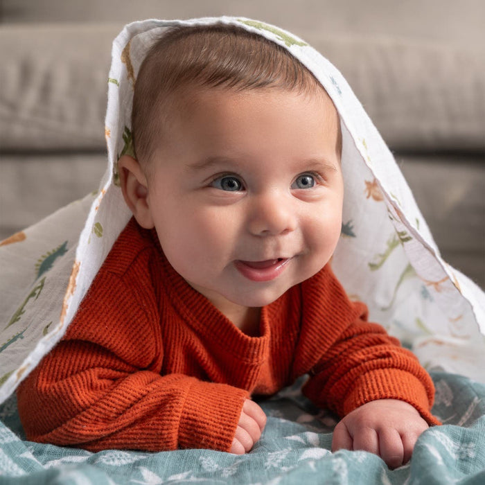A baby with short brown hair lies on a patterned blanket, wearing an orange sweater. An aden + anais essentials cotton muslin swaddle from the 4-pack, featuring colorful prints, drapes over the babys head. The baby smiles forward with a sofa in the background.