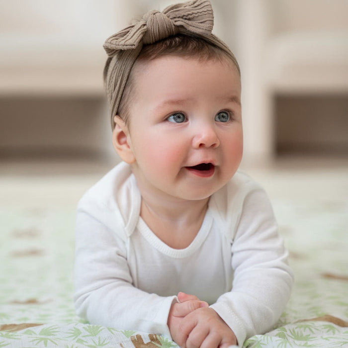 A baby in a white outfit and bow headband lies on their stomach atop an aden + anais essentials cotton muslin swaddle blanket. The baby’s blue eyes and curious expression are highlighted, with hands together, against the softly blurred background of a breathable, light-colored blanket.