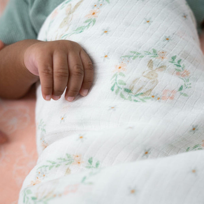A close-up shows a babys hand on an aden + anais essentials cotton muslin swaddle blanket, featuring pastel floral and rabbit designs. The breathable blanket covers a baby in a green garment, resting on a light pink background.