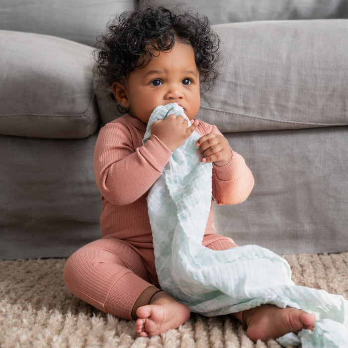 A baby wearing a peach outfit sits on the rug by the sofa, curiously chewing on an aden + anais essentials cotton muslin swaddle blanket. With curly hair and bright eyes, the little one looks adorably inquisitive and content.