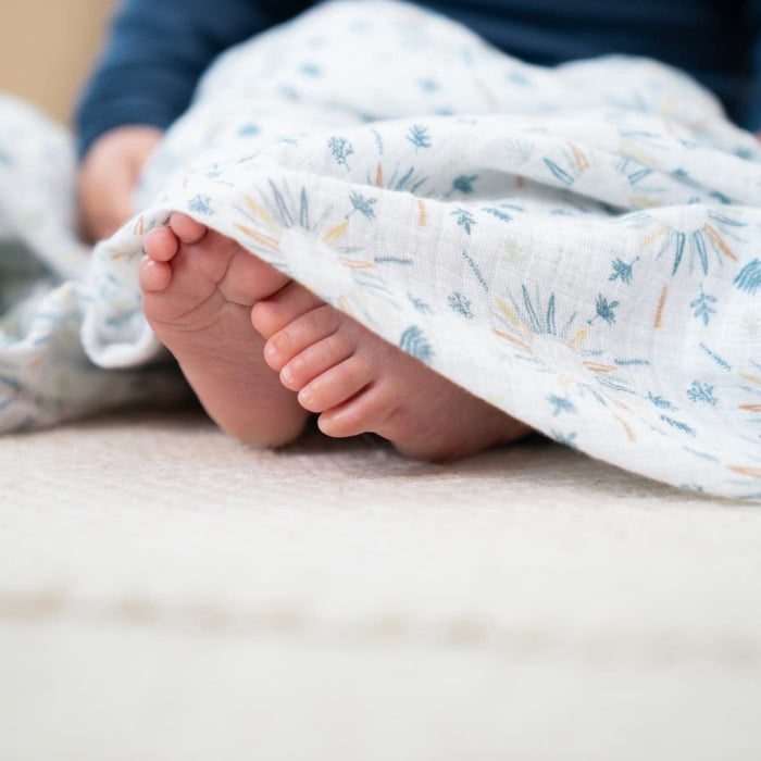 A close-up of a childs feet under an aden + anais essentials cotton muslin swaddle blanket creates a cozy scene. The light, floral-patterned fabric offers comfort as it partially covers the bent legs resting on a textured surface.