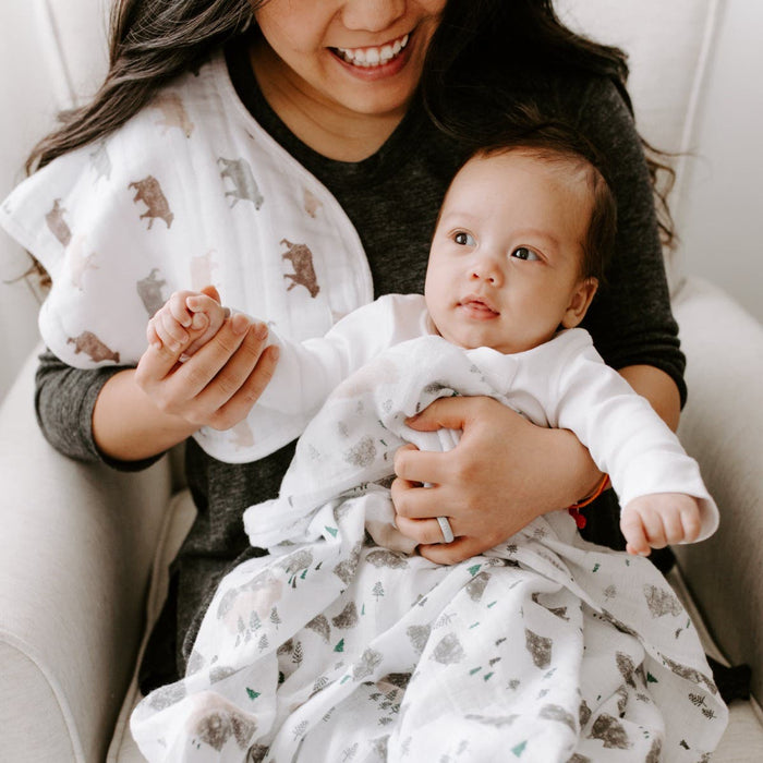 A woman in a dark top holds a baby wrapped in an aden + anais essentials cotton muslin swaddle featuring animal patterns. The baby gazes up as she smiles down. Over her shoulder, a bear-designed cloth from the breathable cotton muslin set showcases its versatility.