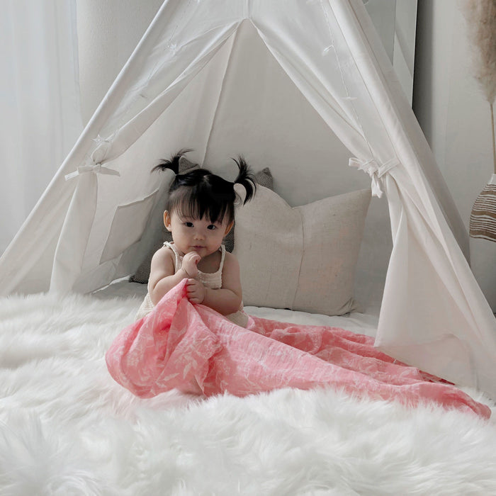 A baby with pigtails sits in a white tent on a fluffy rug, holding a pink aden + anais essentials cotton muslin swaddle blanket. The airy, bright space features simple decor with just a pillow, highlighting the breathable comfort of the cotton muslin fabric.
