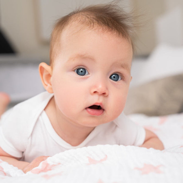 A blue-eyed baby with short light brown hair lies on a white blanket with pink stars, wearing a white onesie and gazing curiously at the camera. Wrapped in an aden + anais Essentials Cotton Muslin Swaddle Blanket, the softly blurred background highlights this versatile baby blanket.