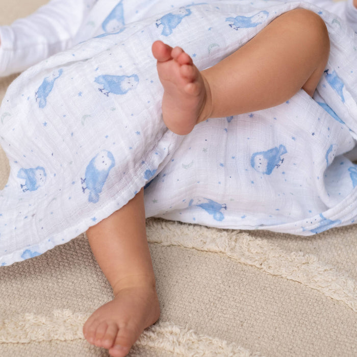 A baby rests on a soft beige surface, partially wrapped in an aden + anais essentials cotton muslin swaddle blanket featuring blue animal prints. One leg playfully extends upward, highlighting comfort and coziness.