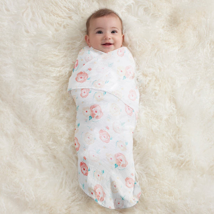 A baby smiles on a cream-colored surface, snugly wrapped in an aden + anais essentials cotton muslin swaddle blanket featuring pink and peach floral patterns.