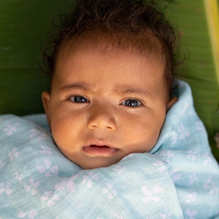 A baby with curly hair lies against a green background, cozily wrapped in an aden + anais essentials cotton muslin swaddle blanket adorned with white floral patterns, gazing wide-eyed and curious.
