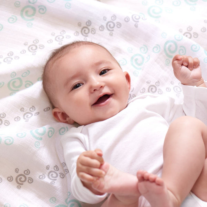 A baby in a white onesie is lying on an aden + anais essentials cotton muslin swaddle blanket, decorated with light blue and gray spirals. The breathable and soft white blanket surrounds the smiling baby, who has arms raised and legs slightly bent.
