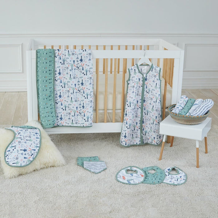 The nursery features a white crib with a breathable aden + anais essentials cotton muslin swaddle blanket and sleep sack. Matching bibs and burp cloths, also in cotton muslin, are on the floor, with extra cloths in a basket on the side table. The room has soft-toned, neutral decor.