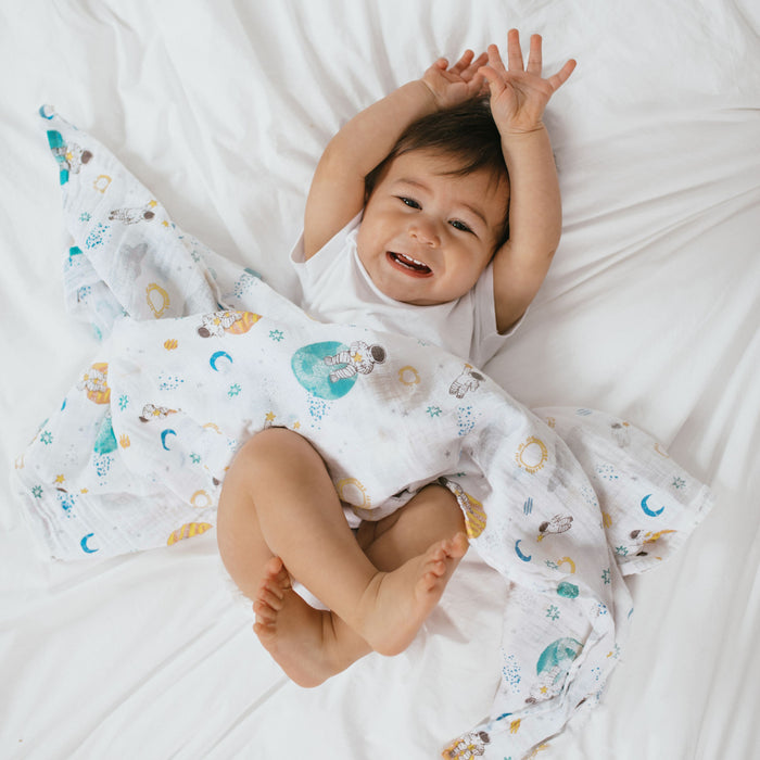 A baby smiles on a white bed, arms raised, wrapped in an aden + anais essentials cotton muslin swaddle blanket featuring colorful stars, moons, and bears.
