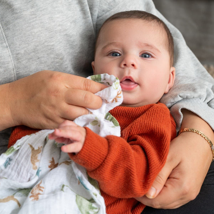 An adult in a gray sweater holds a baby in an orange sweater, gently wiping the babys mouth with an essentials cotton muslin swaddle from aden + anais. The cozy indoor scene highlights parental essentials.