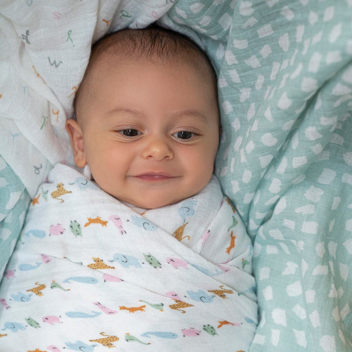 A baby lies serenely on a green blanket with white dots, swaddled in an aden + anais essentials cotton muslin swaddle featuring colorful animal designs. The breathable cotton material keeps the baby calm and comfortable.
